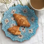 بشقاب مسی رنگ آمیزی شده با رنگ های فیروزه ای و قهوه ای، حاوی کروسان تازه به‌عنوان یک ترکیب زیبا از هنر و خوراکی