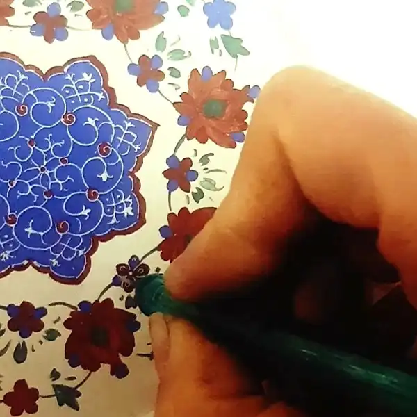 هنرمند (مینا سلیمیان) رنگ های میناکاری را با دقت روی سطح بشقاب نقاشی می کند.