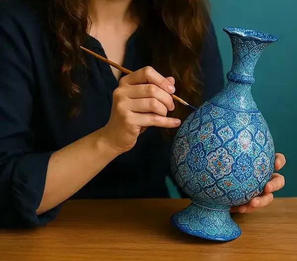 هنرمند ایرانی در حال نقاشی روی گلدان میناکاری با قلم‌موی ظریف در کارگاه سنتی